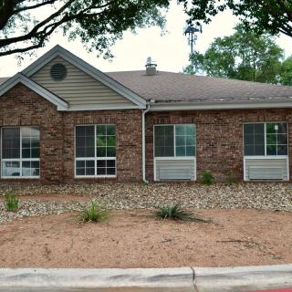 A brick residential building with large windows and minimal landscaping, featuring a row of small plants and trees in the background.