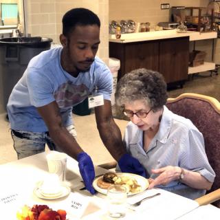 A caregiver assists an elderly woman with her meal at a dining table.