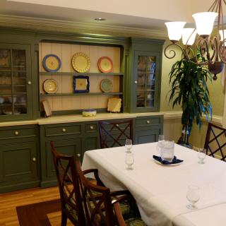 A cozy dining room with a neatly set table, green cabinetry displaying plates, and a decorative plant.