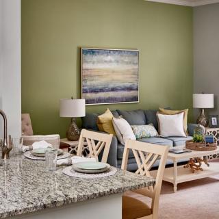 A stylish living room with a green accent wall, featuring a comfortable sofa, tasteful artwork, and a dining area adjacent to a kitchen counter.