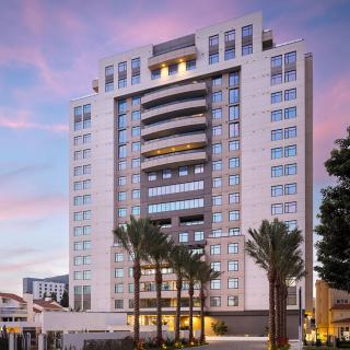 A modern hotel building with palm trees in front, set against a colorful sunset sky.