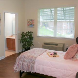 A neatly arranged bedroom with a cozy bed, bedside table, and potted plant beside a large window.