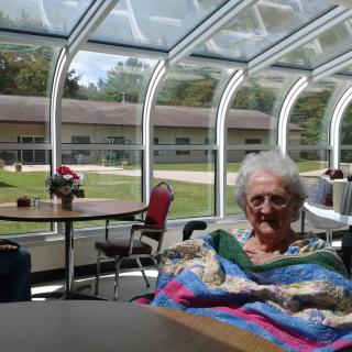 An elderly person sitting indoors under a glass ceiling, wrapped in a colorful blanket.