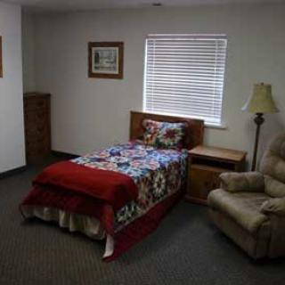 A cozy bedroom featuring a single bed with a colorful quilt, an armchair, and a lamp near the window.