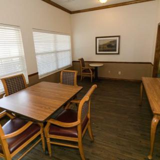 A dining room with wooden tables and chairs, featuring large windows with blinds and a framed artwork on the wall.