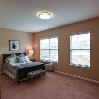 A cozy bedroom with carpeted flooring, featuring a bed, bedside lamp, and two large windows.