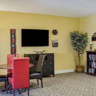 A cozy living and dining area featuring vibrant red chairs, a television mounted on the wall, a decorative clock, an indoor plant, and a bookshelf against yellow walls.