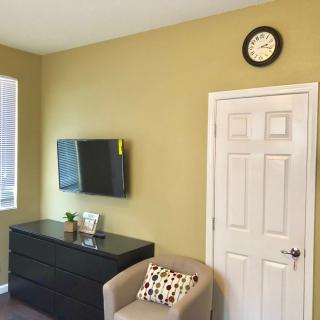 A cozy room interior featuring a wall-mounted television, a black dresser with decorative items, a comfortable chair with a patterned pillow, and a wall clock above the door.