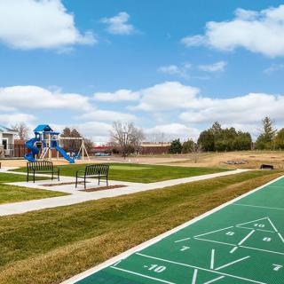 A scenic view of a park featuring a playground, shuffleboard court, benches, and open grassy areas under a blue sky.