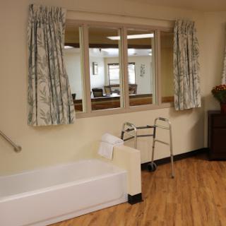 A well-lit healthcare room featuring a bed, walker, and large windows with curtains.