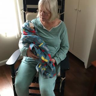 An elderly woman gently holds a baby wrapped in a colorful blanket while sitting in a rocking chair indoors.