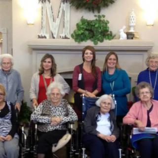 A group of elderly women with caregivers gathered indoors, some seated in wheelchairs.