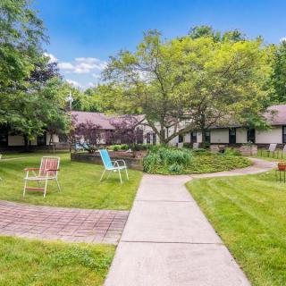 A serene garden setting with chairs and a pathway leading to a building surrounded by lush greenery.