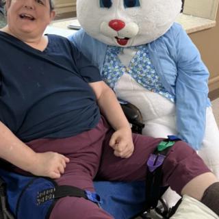 A person in a wheelchair is smiling next to someone dressed as the Easter Bunny indoors.