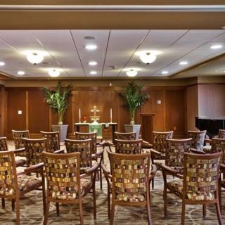 An indoor auditorium with rows of patterned chairs facing a podium, decorated with plants and soft lighting.