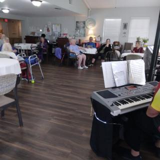 A group of elderly people gathered indoors, enjoying music played on a piano.