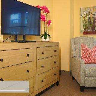 A cozy living room setup featuring a wooden dresser with a television, an armchair, decorative flowers, and wall art.