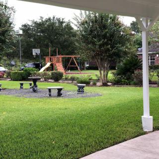 A well-maintained backyard featuring a playground with a swing set and garden benches, surrounded by lush green grass and trees.