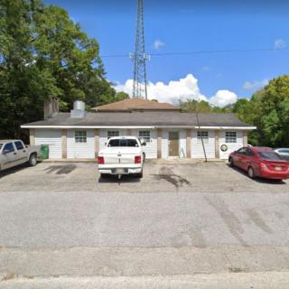 A small building with parked vehicles in front, surrounded by trees and topped with an antenna under a partly cloudy sky.