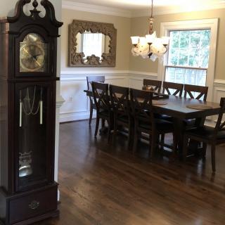 A dining room with a wooden grandfather clock and a large dining table set with chairs, featuring a chandelier and a decorative mirror on the wall.