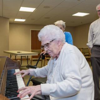 An elderly person playing the piano in a community room with others looking on.