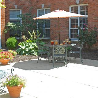 A sunny patio with outdoor furniture and an umbrella, surrounded by plants and a brick building.