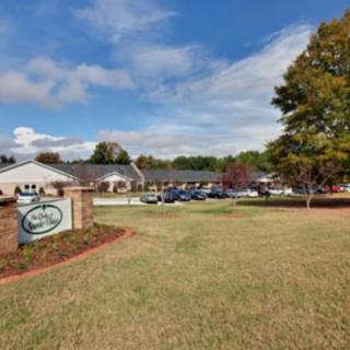 A view of a building with a parking lot, surrounded by trees and grass under a blue sky.