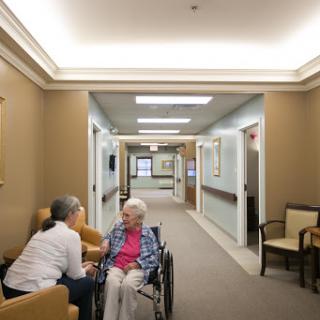 A caregiver attentively interacts with an elderly resident in a well-lit nursing home corridor.