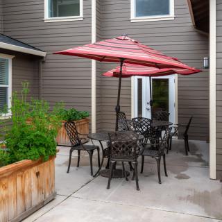 A cozy patio area with a table and chairs under a red umbrella, surrounded by planter boxes and set on a concrete floor.