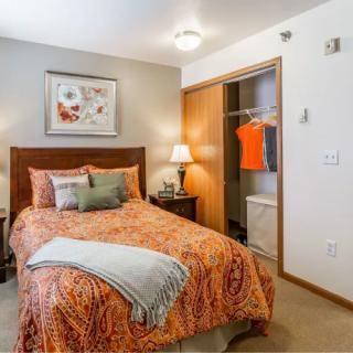 A cozy bedroom featuring a neatly made bed with patterned bedding, a bedside lamp, and an open closet.