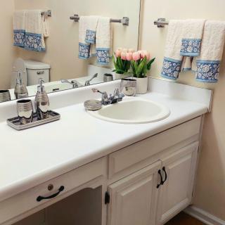 A neatly organized bathroom with a white countertop, sink, mirror, and decorative towels and flowers.