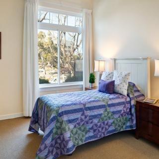 A cozy bedroom with a single bed adorned with colorful bedding, positioned near a large window that lets in natural light. A nightstand with a lamp sits beside the bed.