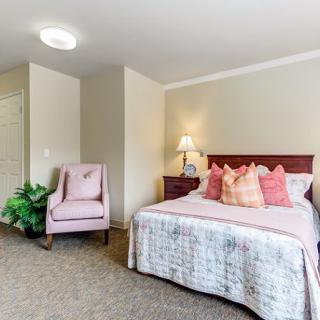 A cozy bedroom with a neatly made bed, a pink chair, and a bedside lamp.
