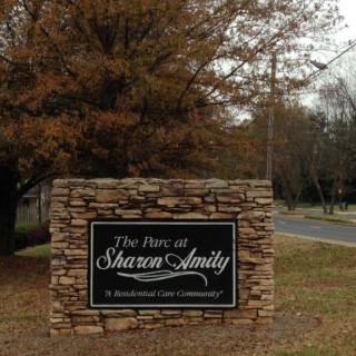 A stone sign marking the entrance to a residential community called 'The Park at Sharon Amity', surrounded by trees and near a road.