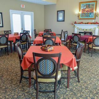 A cozy dining room with several tables covered in red tablecloths, featuring chairs with decorative backs, a carpeted floor, and framed art on the walls.