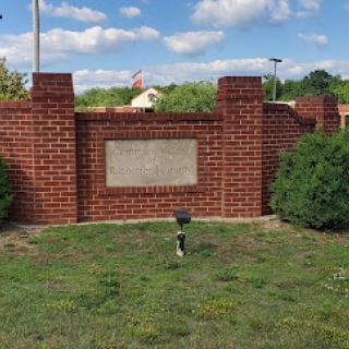 A brick wall with a plaque surrounded by well-maintained grass and bushes under a partly cloudy sky.