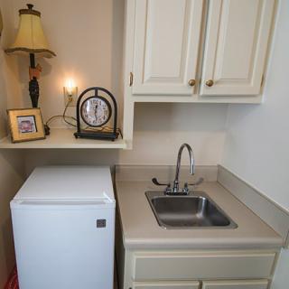 A small kitchen area with a sink, mini fridge, and overhead cabinets.