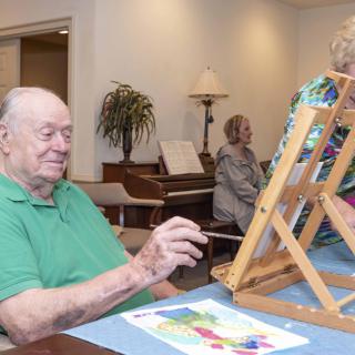 A group of seniors engaging in a painting activity indoors, with one man working on an artwork using an easel.