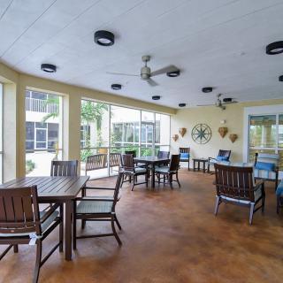 A cozy enclosed patio with multiple tables and chairs arranged for seating, surrounded by large windows and featuring ceiling fans overhead.