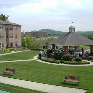 A scenic view of a well-maintained lawn with a gazebo and benches, surrounded by buildings and a group of people gathered nearby.