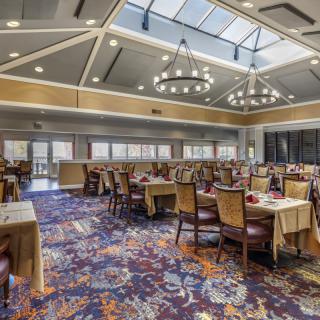 A spacious dining room with elegant chandeliers and a skylight, featuring multiple tables and chairs arranged on a colorful carpet.