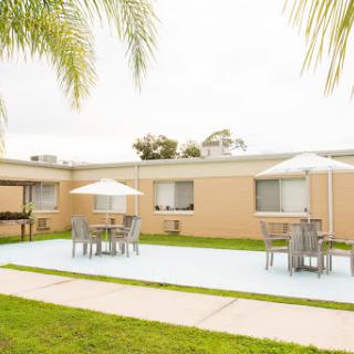 A serene outdoor courtyard featuring tables and chairs under umbrellas, surrounded by a well-maintained lawn and palm trees.