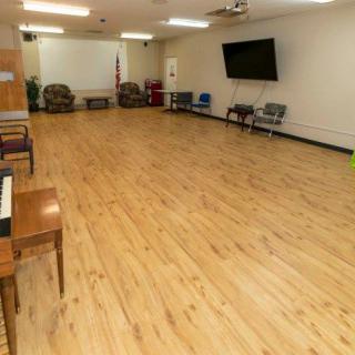 An empty room with wooden floors, a piano on the left, and chairs against the walls.