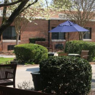 A peaceful courtyard with benches and a blue umbrella, surrounded by manicured bushes and trees.