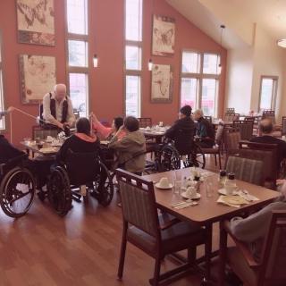 A group of elderly individuals gathered in a dining area, some in wheelchairs, enjoying a meal together.