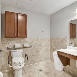 An accessible bathroom with a toilet, sink, and mirror, featuring handrails and tiled walls.