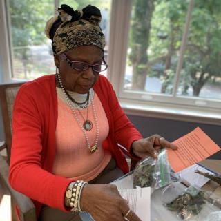 A woman wearing a headscarf and glasses is sitting indoors, reading information from a package.