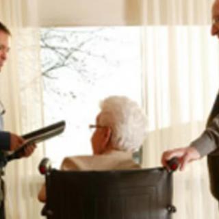 A caregiver and another person engage in a discussion with an elderly person seated in a wheelchair.