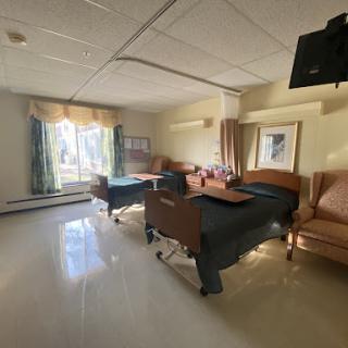 A well-lit hospital room featuring two patient beds, chairs, and a window with a view.