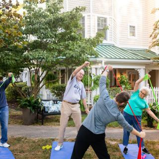 A group of people participating in an outdoor exercise session, performing stretching exercises on yoga mats in a garden setting.